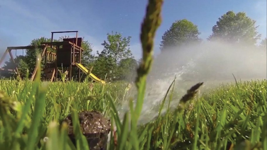 Up close picture of a lawn sprinkler with a small playground with yellow slide in the background