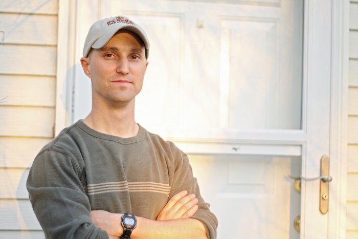 man wearing baseball cap and brown shirt with arms crossed in front of door