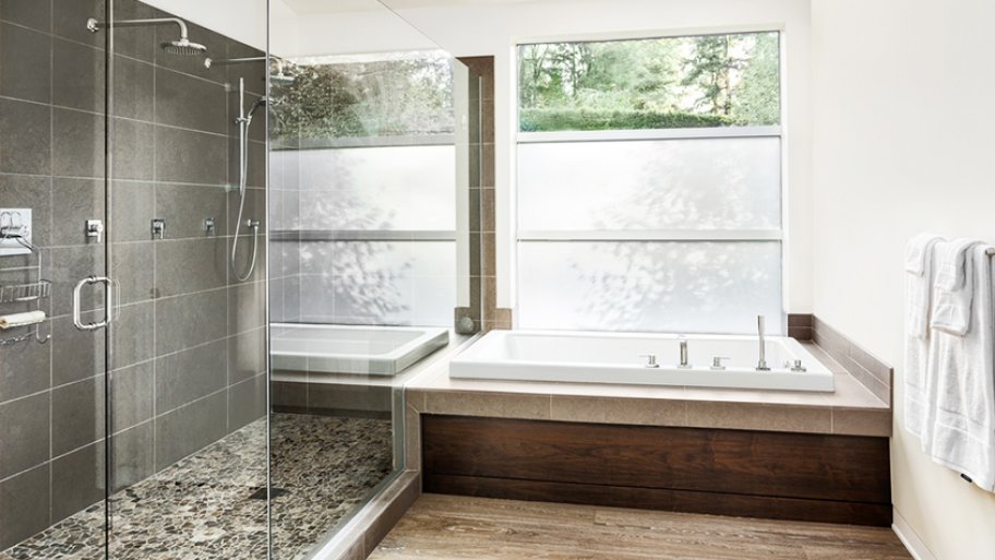 Large glass shower with gray tile, alongside bathtub with wood base