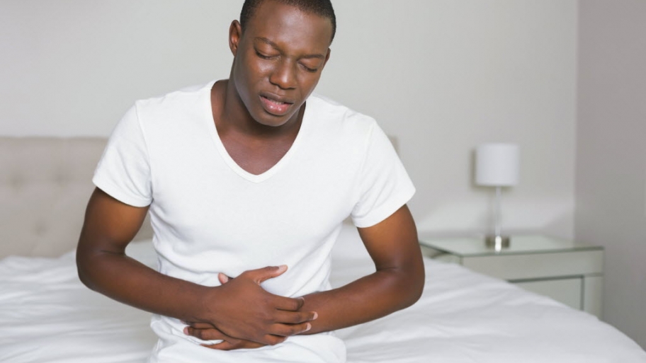 Wincing man sitting on the edge of his bed with a stomach cramp at home in bedroom
