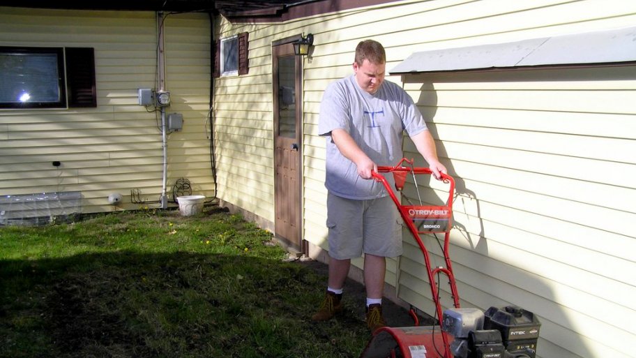 Rototiller Repair Troubleshooting Issues With Garden Tillers Man Pushes A Troy Bilt Bronco Rototiller In The Backyard Alongside His House