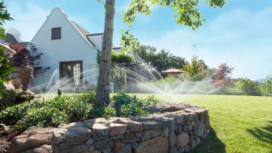 Large yard with multiple sprinklers and a rock ledge with a tree