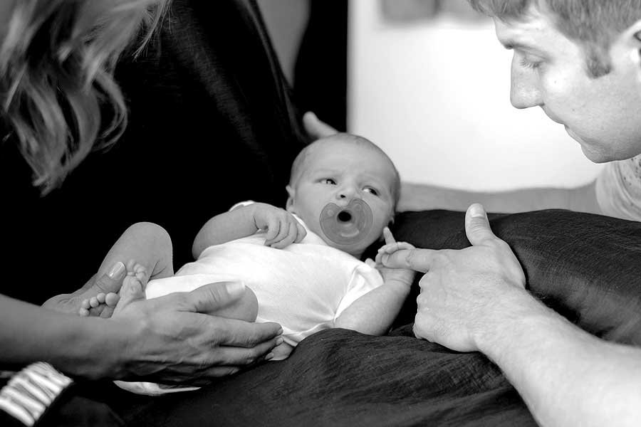 A woman holds a baby while the baby holds a man's finger.